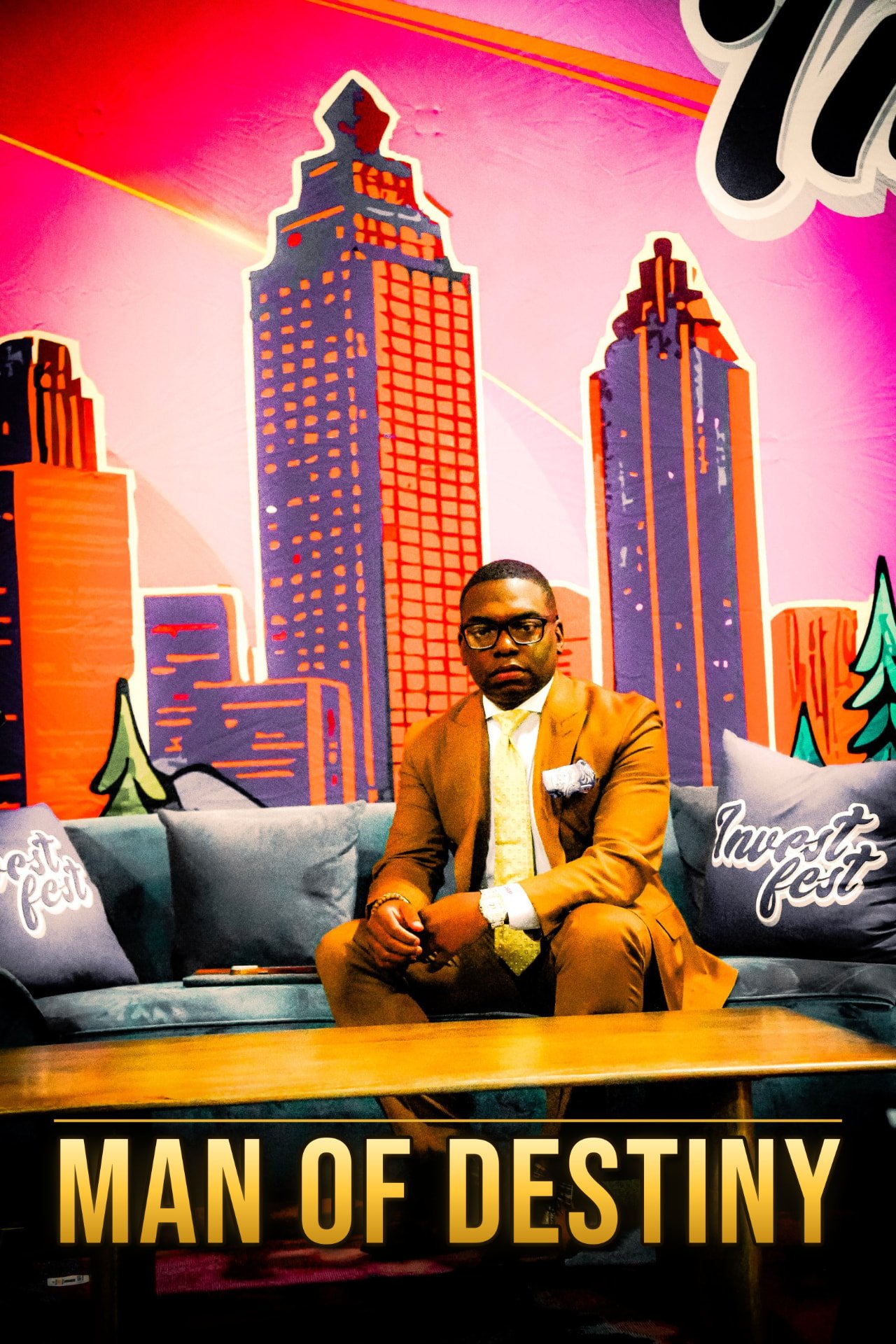 Man in a tan suit seated on a blue couch in front of a skyline mural at Invest Fest 2026, with the text Man of Destiny