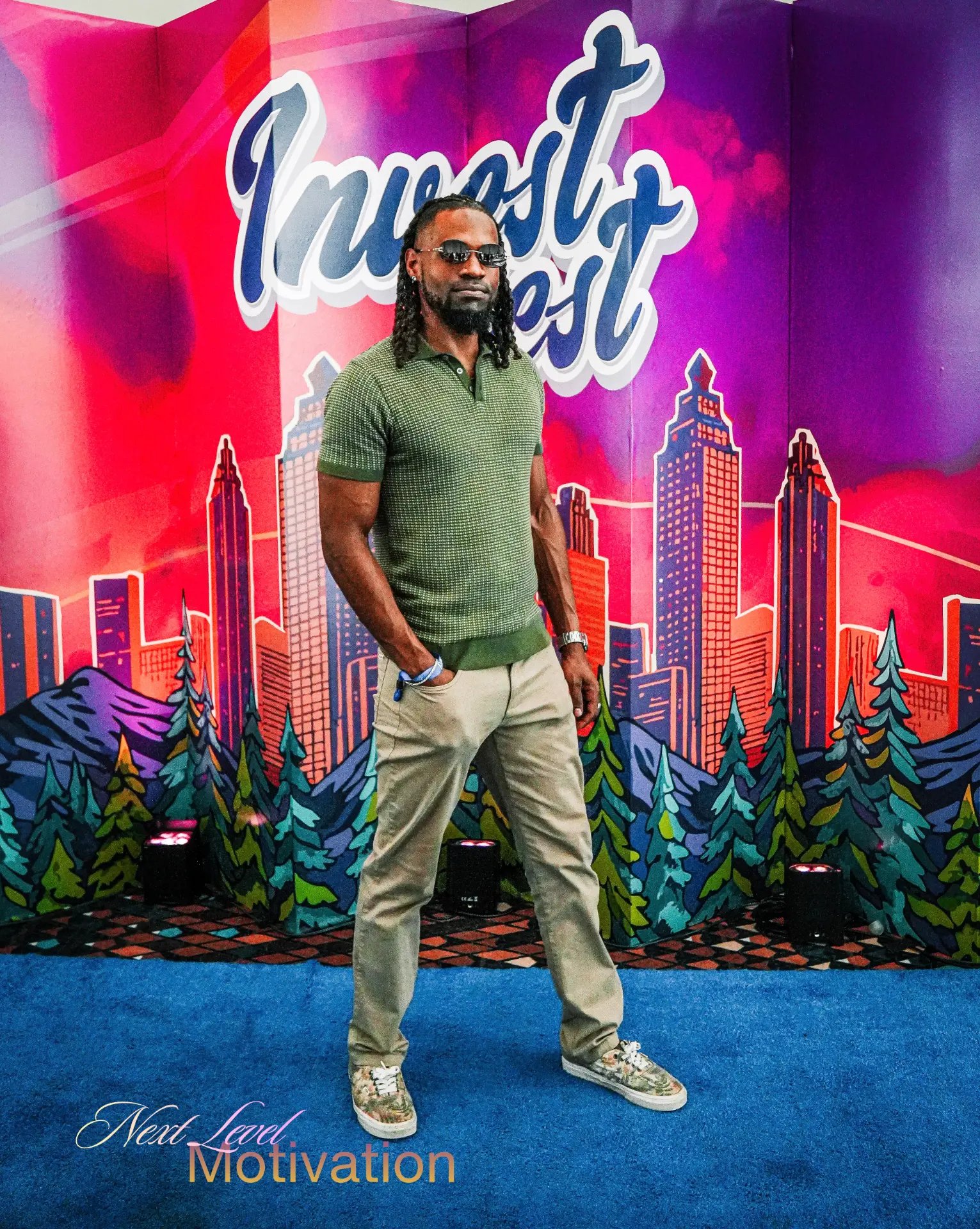 Man with long locs and sunglasses in a green knit polo and khaki pants standing on a blue carpet before a colorful city skyline backdrop