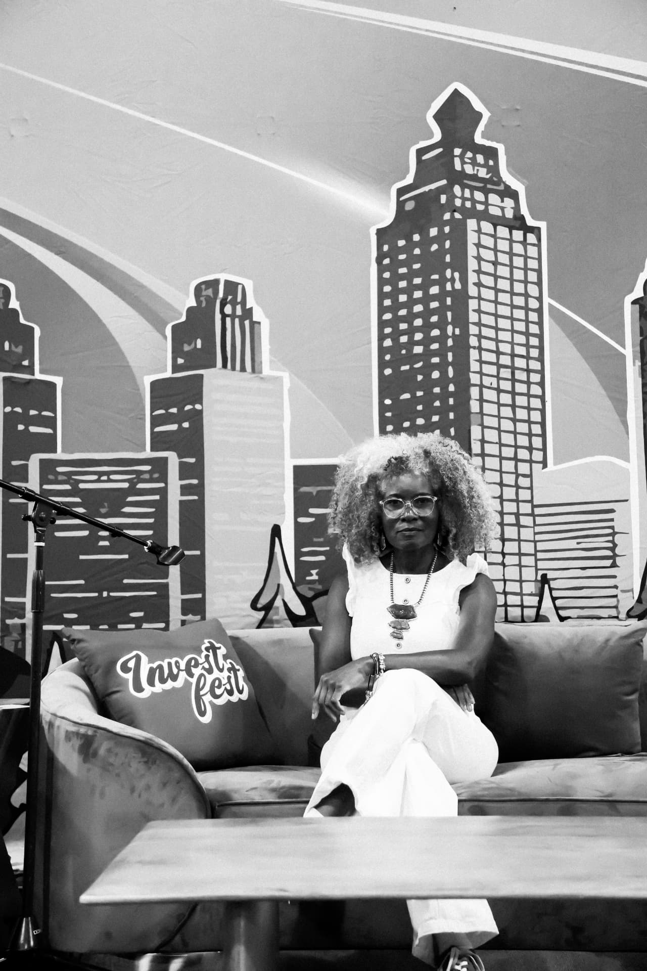 Black and white photo of a woman with curly hair seated on a stage sofa in front of a city skyline mural