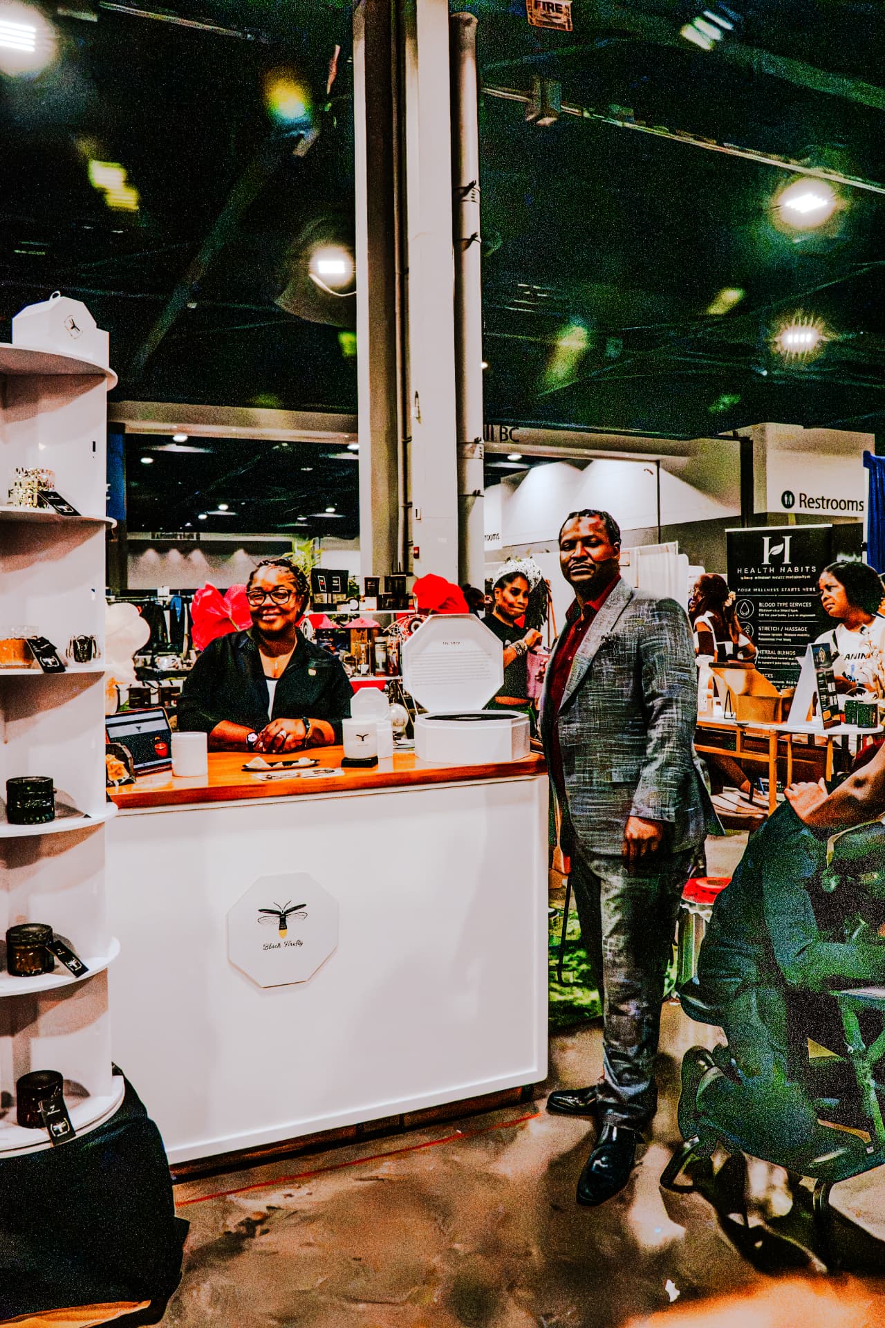 Vendor smiling behind a white candle booth counter while a man in a gray suit stands beside it in the exhibit hall